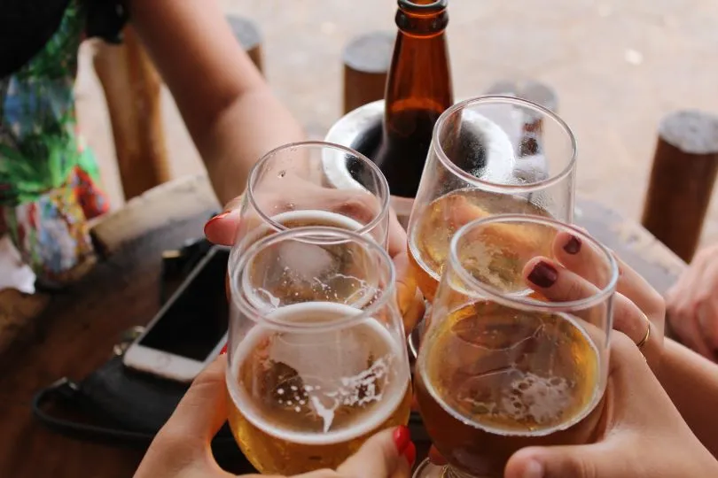 Mehrere Personen stoßen mit Gläsern an, die ein helles, sprudelndes Cider enthalten. Die Szene wirkt gesellig und entspannt, möglicherweise draußen an einem Tisch. Man sieht Hände mit lackierten Fingernägeln, eine Flasche im Hintergrund und ein Smartphone auf dem Tisch, was auf ein lockeres Treffen oder eine Feier hindeutet.