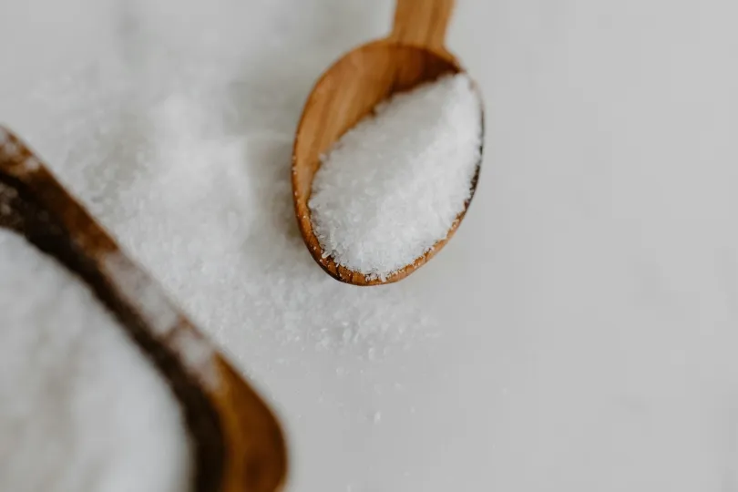 Ein Holzlöffel, gefüllt mit groben, weißen Salzkristallen, liegt auf einer hellen Oberfläche. Ein zweiter Löffel ist unscharf im Hintergrund zu sehen, während einige Salzkörner daneben verstreut sind.