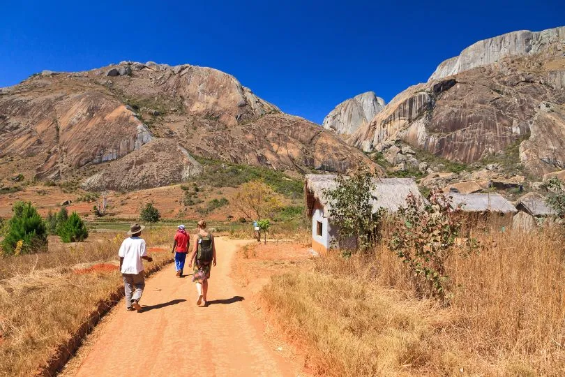 Touristen und ein Einheimischer laufen auf einer trockenen Straße in Madagaskar in Richtung großer Felsen.