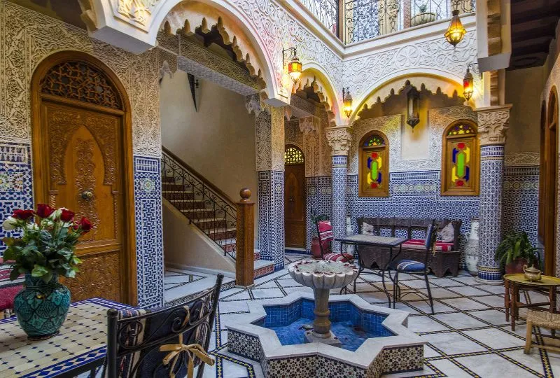 Kunstvolle Riad mit geschnitzten Stuckverzierungen und blau-weißen Zellige-Fliesen sowie einem kleinen Brunnen in der Mitte.