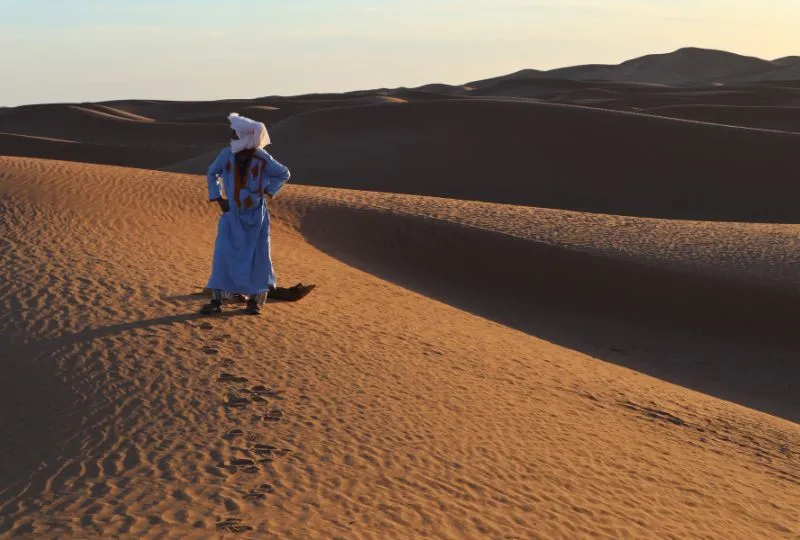 Marokkaner in einem blauen Gewand und weißen Turban steht bei Abendsonne in den Dünen der marokkanischen Wüste.
