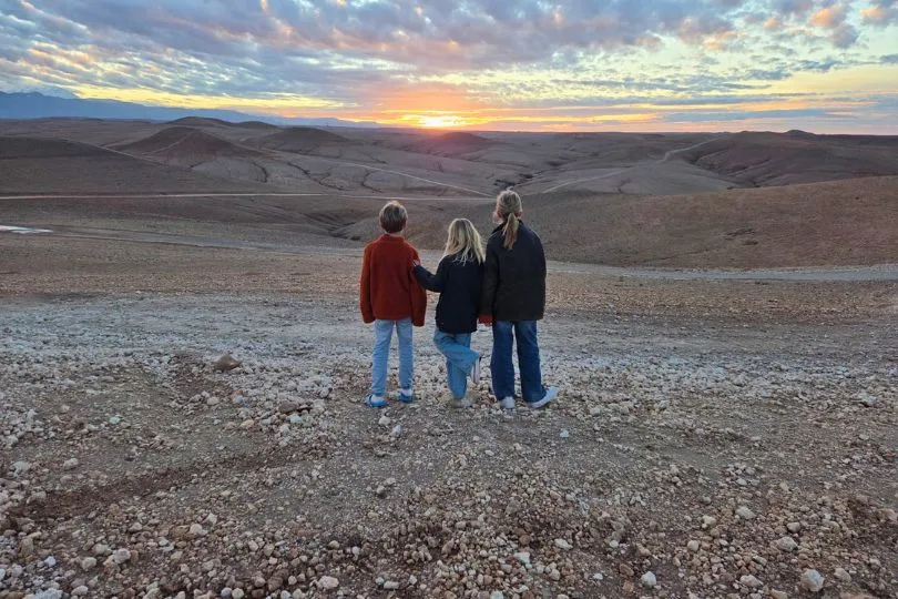 Drei Kinder stehen vor einerkargen Landschaft Marokkos und schauen gemeinsam den Sonnenuntergang.