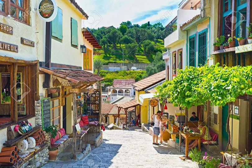 Kleine Gasse in Montenegro mit hellen Häusern, Menschen im Café und bunten Produkten zum Verkauf.