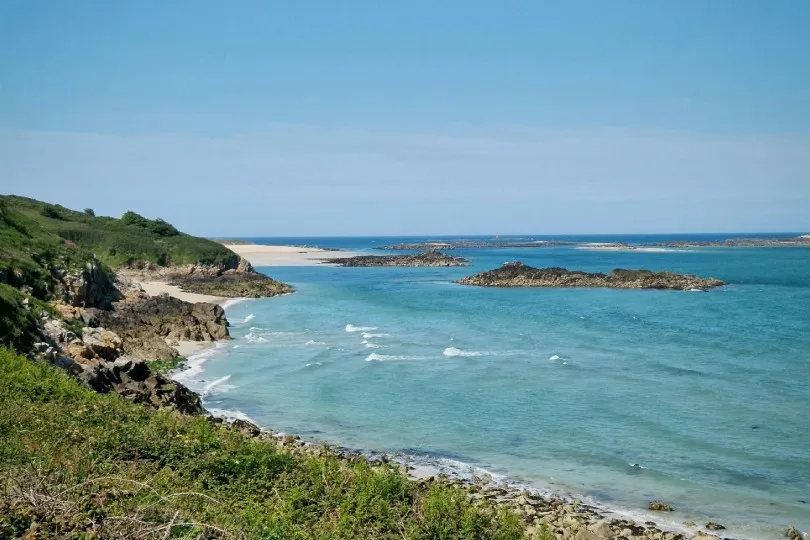 Küstenlandschaft mit klarem Wasser, kleinen Wellen und felsigen Inseln, links ein grüner Hang und im Hintergrund ein heller Sandstrand unter blauem Himmel.