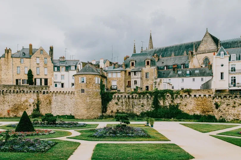 Historische Stadt mit steinernen Häusern und einer alten Stadtmauer, davor ein gepflegter Garten mit Wegen und Beeten unter einem bewölkten Himmel.