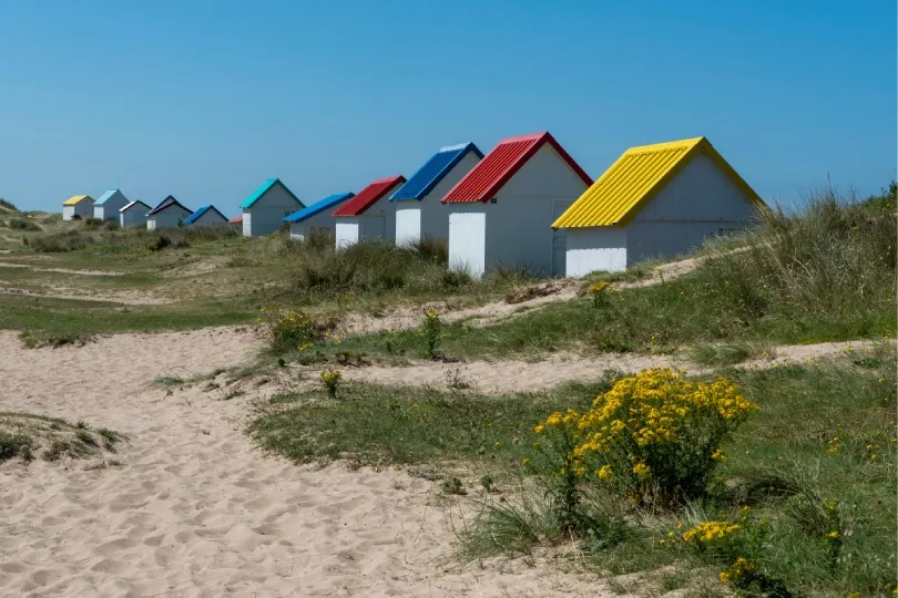 **Alt-Text auf Deutsch:**  **Eine Reihe kleiner weißer Strandhütten mit bunten Dächern steht zwischen Sanddünen und Gras, unter einem klaren blauen Himmel.**