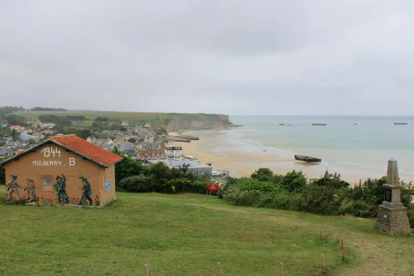 Blick über Arromanches-les-Bains mit Strand, Meer und Resten des Mulberry-Hafens vor der Küste. Im Vordergrund stehen ein Gebäude mit Wandbild zur Landung 1944 und ein kleines Denkmal auf einer Wiese.
