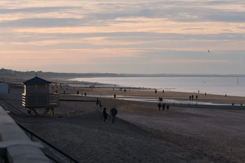 Menschen spazieren bei gedämpftem Abendlicht über einen weiten Sandstrand, neben einem Strandhäuschen mit Blick auf das ruhige Meer.