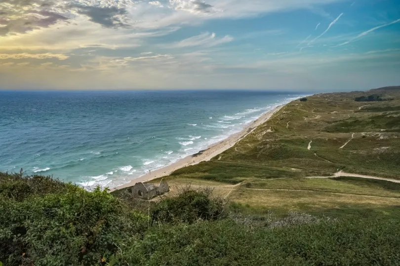 Blick von den Dünen d’Hatainville auf einen langen Sandstrand, türkisblaues Meer und weite grüne Küstenlandschaft unter dramatischem Himmel.