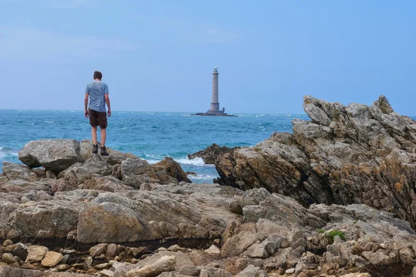 Ein Mann steht auf Felsen an der Küste und blickt über das Meer zu einem Leuchtturm auf einer kleinen vorgelagerten Insel.