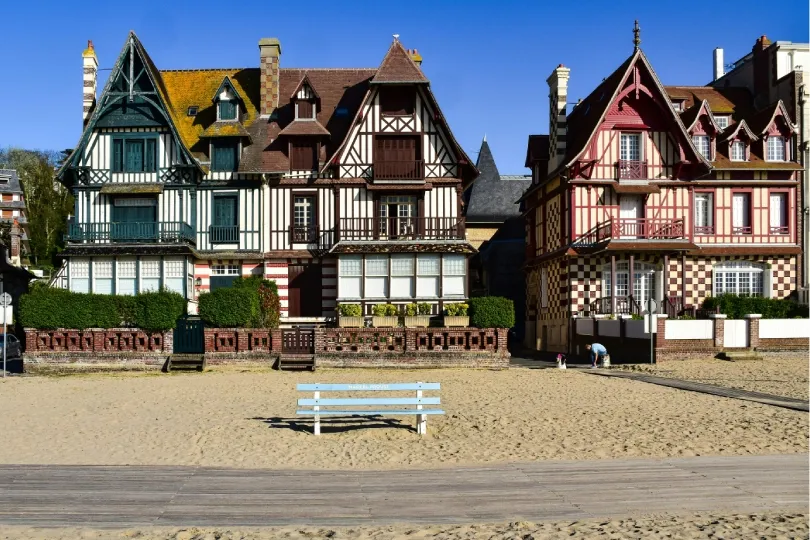 Fachwerkhäuser mit farbigen Holzfassaden stehen direkt hinter einem Sandstrand in Deauville. Im Vordergrund sind eine Holzpromenade und eine leere hellblaue Bank zu sehen.