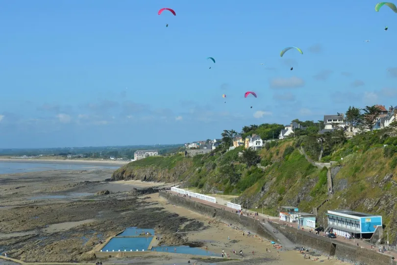 **Blick auf eine felsige Küste mit Strandpromenade und Häusern am Hang, während mehrere bunte Gleitschirme über dem Meer und der Küstenlandschaft fliegen.**