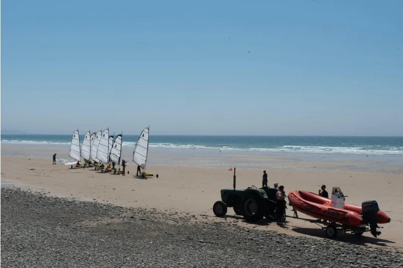 An einem weiten Sandstrand stehen mehrere Strandsegler mit weißen Segeln nahe am Wasser, während rechts ein Traktor ein rotes Schlauchboot auf einem Anhänger zieht.