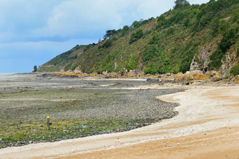 **Weiter Sandstrand bei Ebbe mit felsigen Abschnitten und einer grünen Steilküste, auf deren Hang einzelne Häuser stehen.**