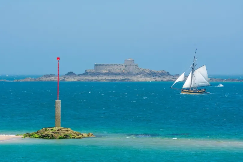 Ein Segelboot fährt über türkisblaues Meer vor einer felsigen Insel mit einer Festung; im Vordergrund steht eine rote Seezeichen-Stange im flachen Wasser.