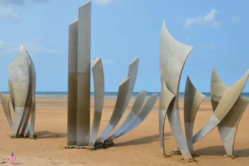 **Alt-Text:** Mehrere hohe, abstrakte Metallskulpturen stehen auf einem breiten Sandstrand vor dem Meer. Im Vordergrund ist eine kleine US-Flagge im Sand zu sehen, darüber ein blauer Himmel mit wenigen Wolken.
