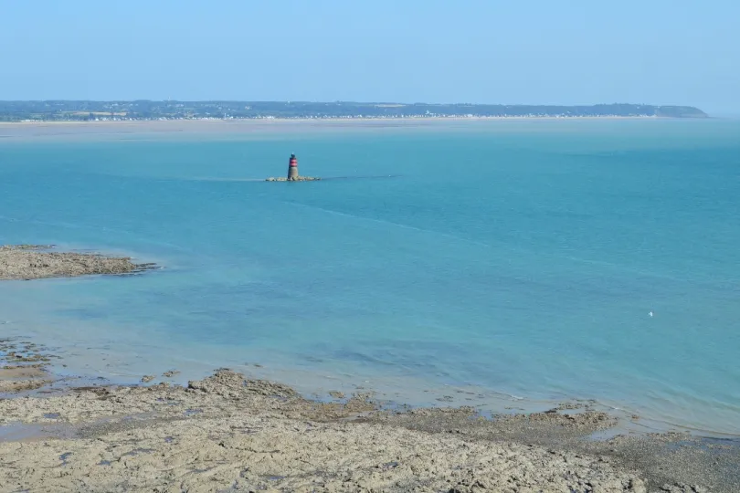 Blick über türkisblaues Meer mit felsiger Küste im Vordergrund. In der Ferne steht ein kleiner rot-schwarzer Leuchtturm auf einer Felseninsel, dahinter ist eine flache Küstenlandschaft zu sehen.