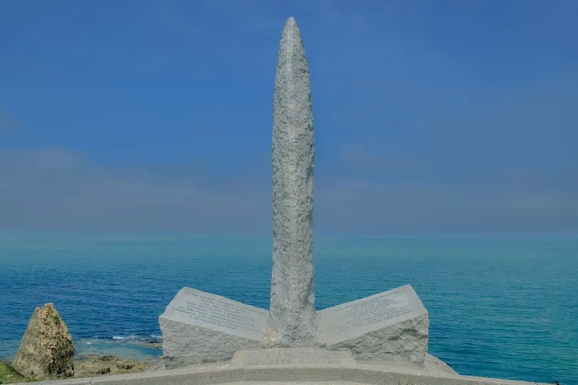 Ein steinernes Denkmal mit hohem, schmalem Obelisken steht auf einer Klippe vor dem blauen Meer. Links sind Felsen und die Küste zu sehen, im Hintergrund ein klarer Himmel.