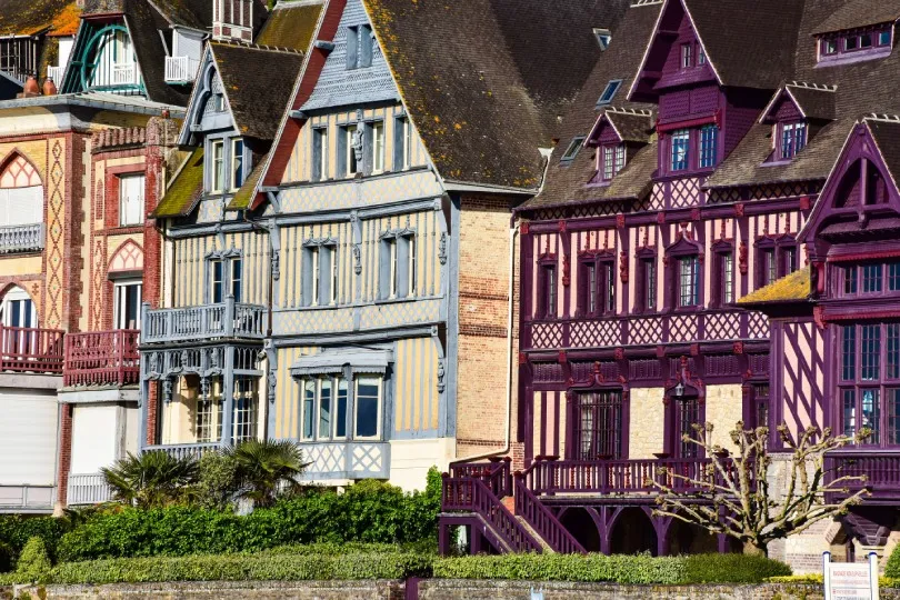 Bunte Fachwerkhäuser mit steilen Dächern stehen dicht nebeneinander. Die Fassaden sind in Pastelltönen und kräftigem Violett gestrichen, davor befinden sich Hecken, Balkone und ein kleiner Baum.