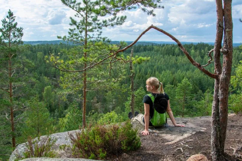 Wanderin sitzt auf einem Felsen mit Aussicht über einen dichten Nadelwald in Finnland.