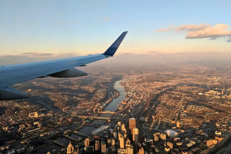 Flugzeugflügel mit Aussicht auf eine große Stadt mit Fluss beim Sonnenuntergang.