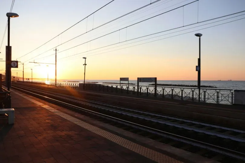 Untergehende Sonne strahlt über die Gleise an einer Zugstation, direkt vor dem Wasser.