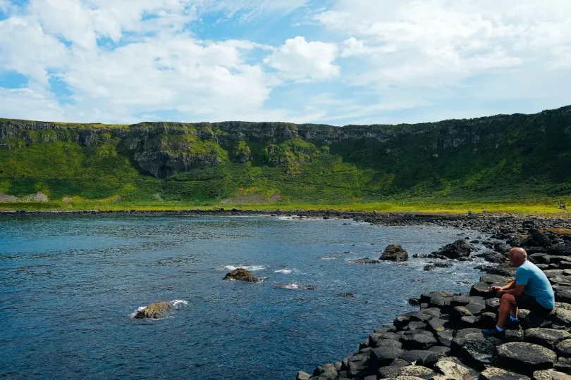 Ein Mann sitzt in einer Bucht in Irland und beobachtet das Meer. Im Hintergrund sind bewachsene Feldwände zu sehen.