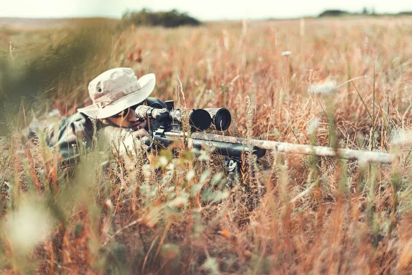 Jäger mit Tarnausrüstung sitzt in einem Feld und schaut durch sein Gewehr, bereit zum Abschuss.