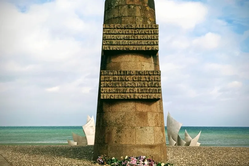 Steinernes Denkmal am Strand von Lion-sur-Mer mit Inschriften zu den alliierten Streitkräften. Im Hintergrund liegen Meer und moderne Gedenkskulpturen unter leicht bewölktem Himmel.