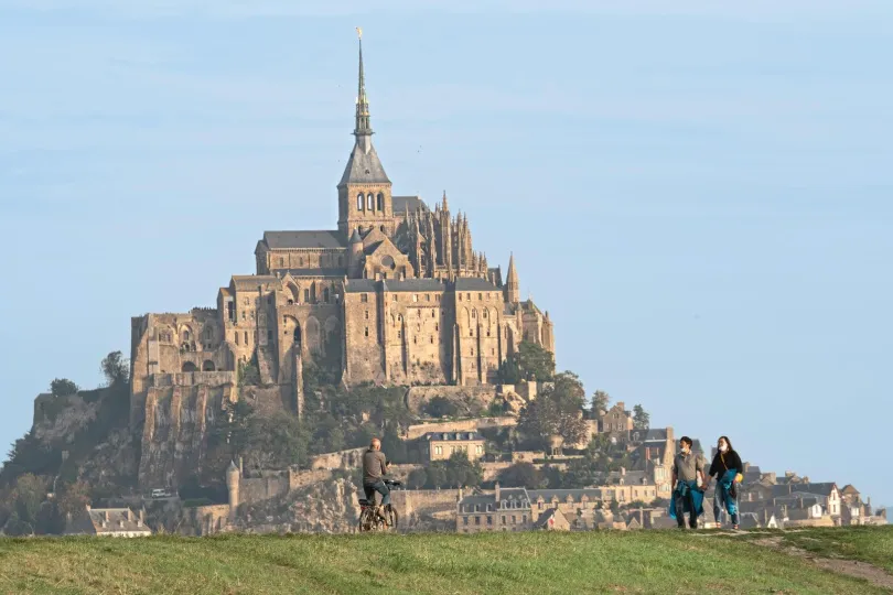 Blick auf den Mont-Saint-Michel mit seiner Abtei im Hintergrund, während Menschen im Vordergrund über eine grüne Wiese gehen und Rad fahren.