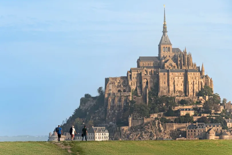Drei Personen gehen über eine grüne Wiese auf den Mont-Saint-Michel mit seiner markanten Abtei zu.