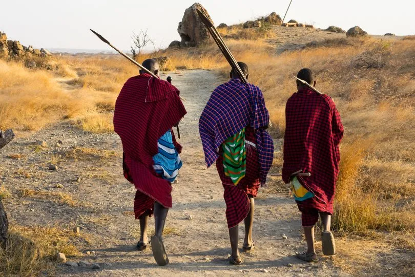 Drei Maasai in traditionellen Gewändern laufen durch die Steppenlandschaft Namibias.