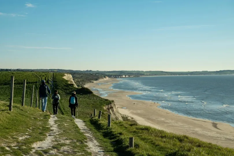 Alt-Text: Drei Wanderer gehen auf einem Küstenpfad entlang grüner Klippen mit Blick auf einen weiten Sandstrand und das Meer in der Normandie.