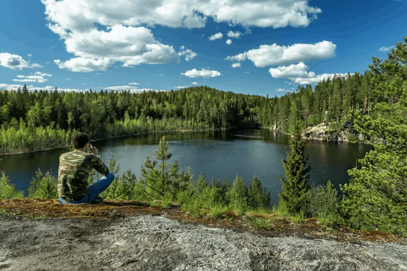 Ein Mann sitzt auf einem Felsen und blickt über einen stillen See, umgeben von dichtem grünem Wald unter einem blauen Himmel mit weißen Wolken.