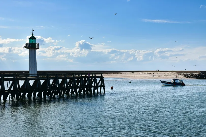 Leuchtturm auf einer Holzmole am Hafen von Honfleur, dahinter Sandbank und Meer. Ein kleines Boot fährt über das Wasser, während Möwen am blauen Himmel fliegen.