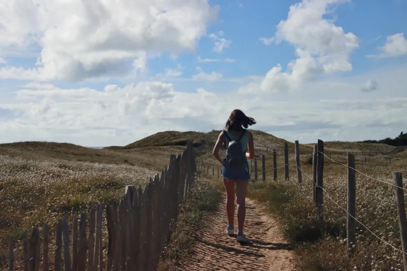 Wanderin mit Rucksack läuft auf einem sandigen Pfad zwischen Dünen und Holzzäunen unter wolkigem Himmel.