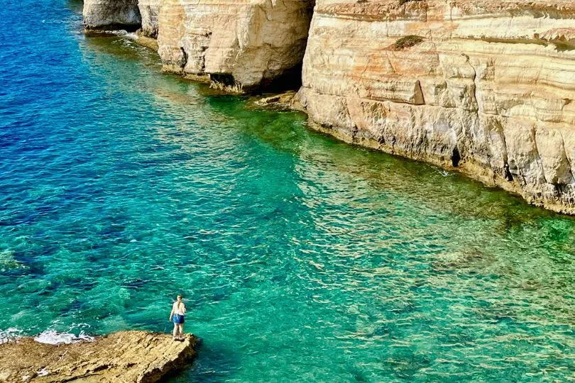 Frau steht auf einem Felsen an einer Bucht in Zypern mit türkisem Wasser und hohen ockerfarbigen Felsen.