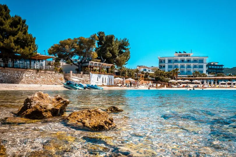 Strand bei Ksamil in Albanien mit klarem Wasser und einigen kleinen Booten und Gebäuden im Hintergrund.