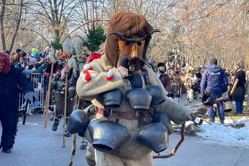 Person in einer Holzmaske mit Kuhglocken um den Hals (ein Kukeri) läuft bei einer Parade über die Straße in Bulgarien.