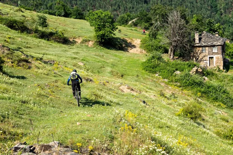 Mountainbiker fährt über eine Wiese vorbei an einer kleinen Steinhütte in den Bergen Bulgariens.