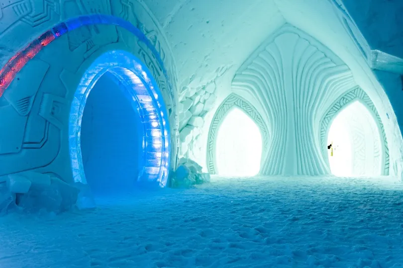 Innenansicht eines kunstvoll gestalteten Eishotels in Schwedisch-Lappland mit blau beleuchtetem Rundbogen, gewölbten Eiswänden und zwei hellen, spitzbogigen Durchgängen.