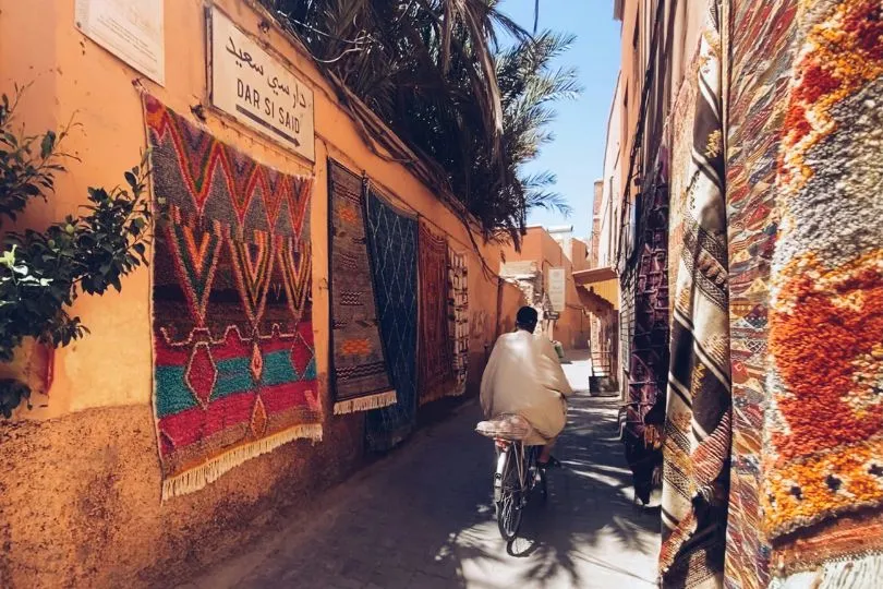 Ein Marokkaner fährt durch eine kleine Gasse in Marokko. An den Wändern hängen bunte Teppiche.