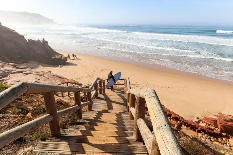 Ein Mann mit Surfbrett steht am Ende einer Brücke auf einem Sandstrand in Portugal.
