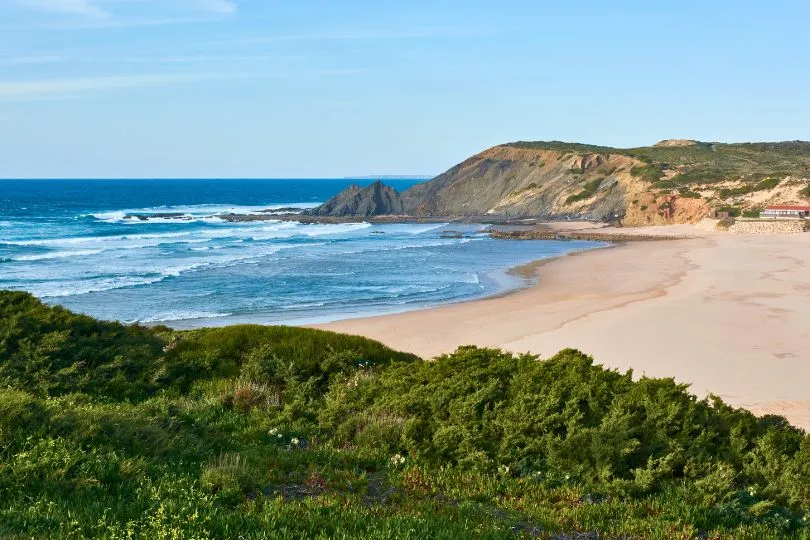 Der Paraia da Amoreira in Portugal mit seinem weißen Strand, üppiger Vegetation im Vordergrund und einer Klippe im Hintergrund. 