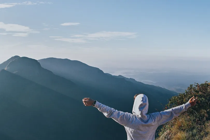 Eine Person in einem hellgrauen Kapuzenpullover steht mit ausgebreiteten Armen auf einem Berggipfel und blickt über mehrere sanft geschichtete Bergketten.