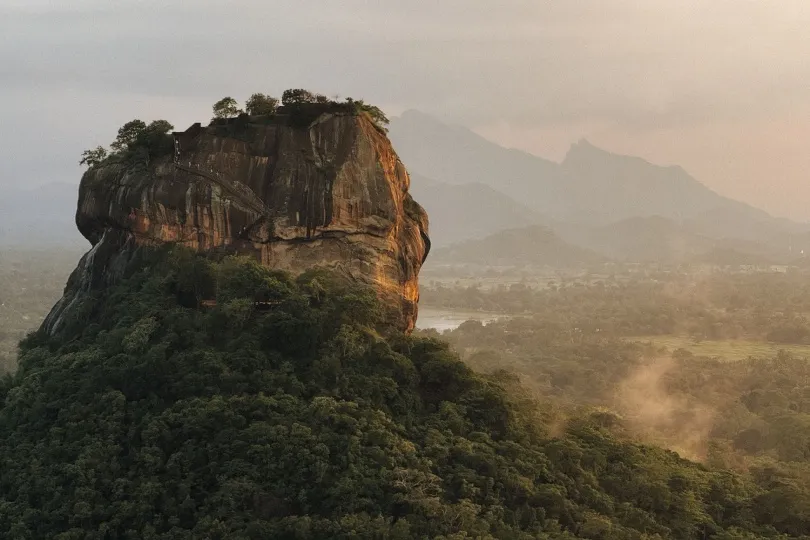 Ein großer, steiler Felsen ragt aus dichtem, grünem Wald empor; im Hintergrund liegen neblige Berge und eine weite Landschaft im warmen Morgenlicht.