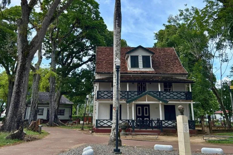 Typisches weißes Holzhaus mit Balkonen und rotem Dach steht zwischen Bäumen in Paramaribo, der Hauptstadt Surinames.