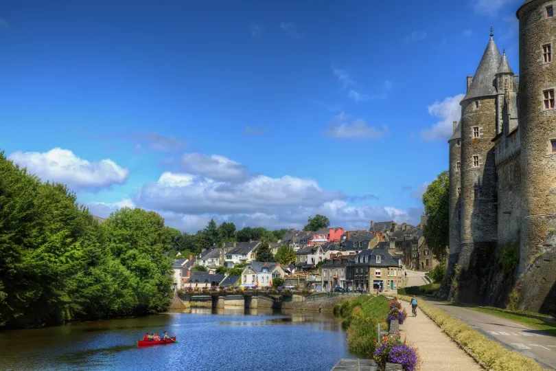 Blick auf Josselin in der Bretagne mit dem imposanten Château de Josselin, dem Fluss Oust, einer kleinen Brücke und einem roten Boot auf dem Wasser.