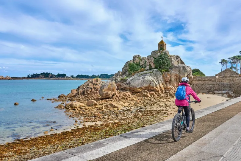 Radfahrerin an der bretonischen Küste bei Penvénan mit felsiger Kapelle, Sandstrand und türkisblauem Meer.