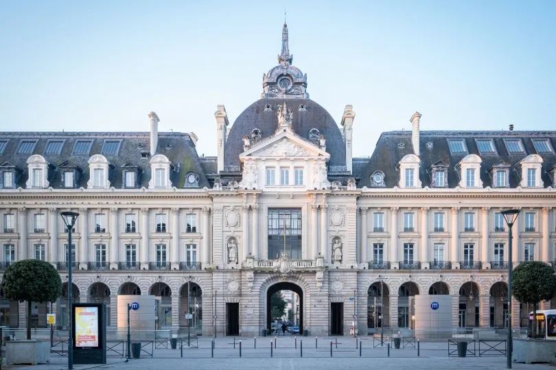 Prächtiges historisches Gebäude in Rennes mit heller Steinfassade, zahlreichen Fenstern und einem hohen, reich verzierten Kuppeldach. Im Erdgeschoss führen Arkaden und ein großes Tor durch das symmetrisch gestaltete Bauwerk.